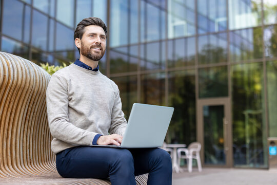 A smiling young man sits on a bench on the street near an office building, holds a laptop in his lap and looks thoughtfully ahead