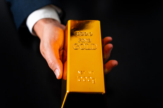 Gold investment secures financial savings. Gold remains strong during market volatility. Businessman carries gold confidently with hands. Gold bar represents stable capital.