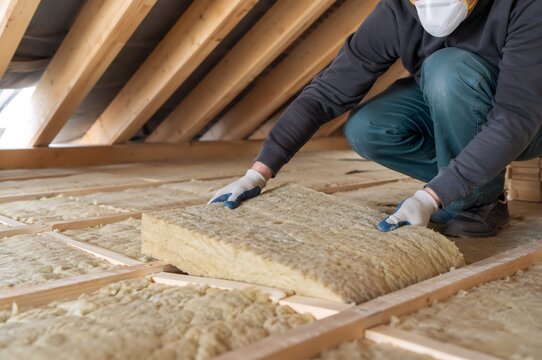 Worker installing thermal insulation between wooden roof beams in an attic. Home renovation and energy efficiency preparation for cold winter weather, with protective equipment and copy space.