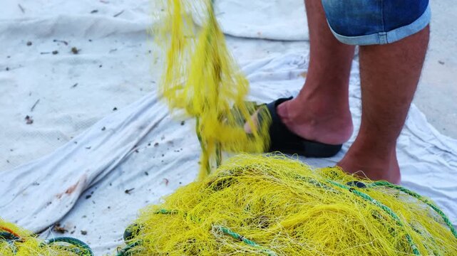 A low-angle close-up shot of a nylon fishing net falling in continuous motion and stacking into a neat pile at the fisherman's feet on the dock. Bright daylight highlights the intricate textures of th
