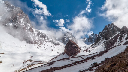 Broad snowy valley framed by rocky peaks and drifting clouds at Bogdanovich Glacier in the Ile Alatau mountain range. © Kolchanov