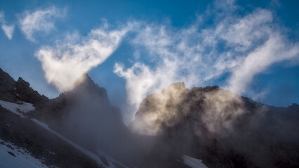 Sunlight breaks through drifting clouds above steep rocky walls and melting snowfields creating a dramatic alpine atmosphere at Bogdanovich Glacier. © Kolchanov