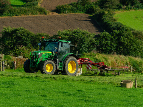 West Cork, Ireland, August 25, 2023. A green tractor tows a red tedder across a grassy field. The machine is currently engaged in tilling the land. Irish countryside is characterised by lush grass.