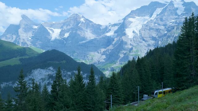 Yellow train traveling on a railway through the mountains of switzerland with peaks in the background. Switzerland