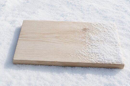 Wooden plank resting on a snow-covered surface with a light dusting of snow on one edge, showcasing natural wood grain and texture in a winter setting