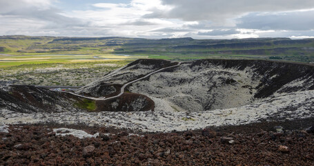 A scenic view of Grabrok volcanic crater with a wooden trail, dark volcanic rock, moss and green countryside under a dramatic cloudy sky in Iceland. © Asanee