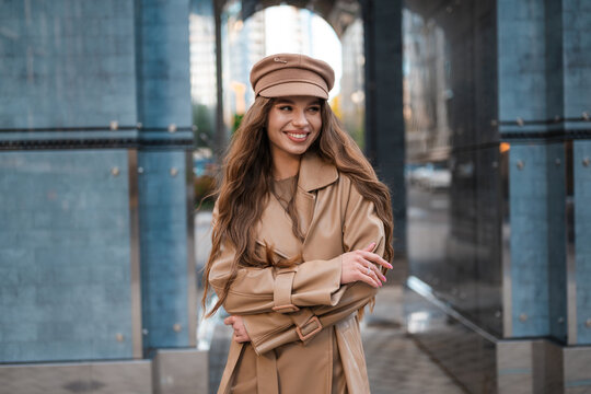 Stylish woman in beige trench coat and cap smiles outdoors in modern city setting, framed by glass pillars, arches, and clean architectural lines under bright daylight