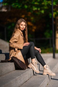Stylish young woman seated on concrete steps outdoors in warm sunlight, wearing tan trench coat, beige boots, and black pants beside green park surroundings, with woman, female, portrait, fashion
