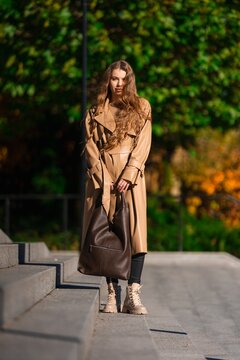 Woman in beige trench coat stands on sunlit concrete steps outdoors, carrying large brown leather bag, styled for urban autumn fashion with warm golden light, with woman, trench, coat, beige detail