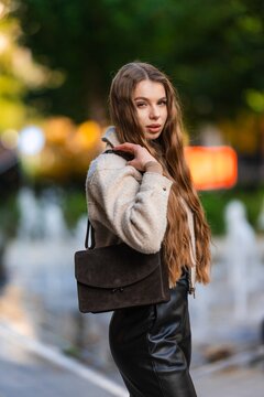 Portrait of young woman with long light brown hair wearing cream beige coat and dark leather crossbody bag outdoors in sunny urban park with soft blurred background