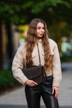 Stylish young woman in beige coat with long wavy brunette hair holding dark handbag while standing outdoors in city park during autumn daylight fashion portrait