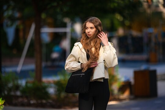 Elegant woman in beige trench coat standing outdoors on city street with soft sunlight, wavy brown hair, confident pose and chic fashion style against blurred urban background