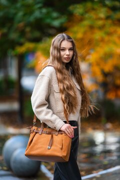 Young woman with long wavy brown hair wearing beige coat and tan leather handbag standing outdoors in autumn park with colorful orange and yellow leaves, with woman, outdoor, autumn, fall detail
