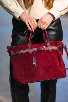 Close view of hands with rings resting on burgundy suede handbag held by woman in beige jacket, showing textured straps, silver hardware, soft outdoor urban background