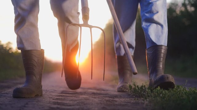 Farmer boots pitchforks road. A man and woman walk together on a dirt path, each holding a farming tool. The man and woman walk towards the sunset, dust rising from the ground.