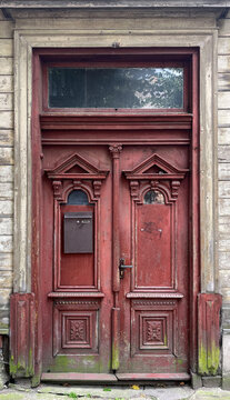 Ornate historic wooden double door with carved decorative panels, transom window, and a mounted mailbox, showing weathered red paint, aged timber, and traditional architectural craftsmanship.