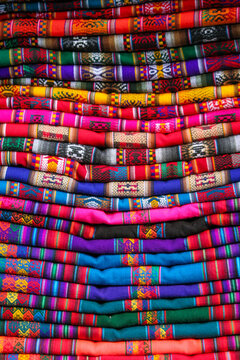 A stack of hand-woven textiles featuring vibrant colors and traditional geometric patterns is neatly piled. These artisanal fabrics reflect rich Andean craftsmanship.