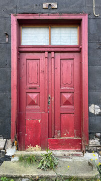 Weathered red wooden double door set within a dark aged fa&ccedil;ade, featuring peeling paint, geometric panels, a transom window, and signs of decay that reflect historic architectural character.
