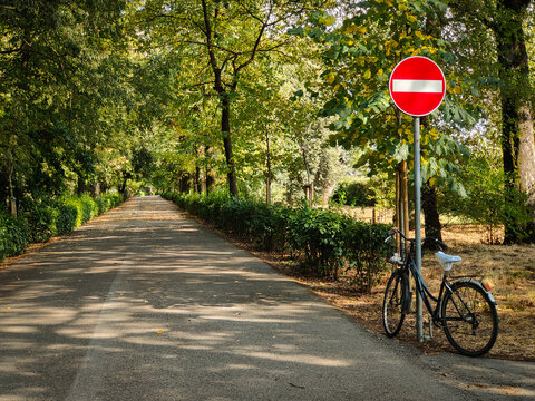 A bicycle is parked against a metal post topped with a red no entry traffic sign on the side of a peaceful, tree-lined path in a park on a bright sunny day.