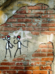 A textured brick wall features rustic street art depicting two stick figures with red hats holding tools. This simple graffiti is painted on a patch of weathered plaster. © Andrea