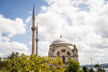 Exterior view of Ortakoy Mosque featuring minarets and central dome framed by green trees under cloudy sky. The image represents Islamic architecture, cultural heritage, and tourism in Turkey. © Tetiana