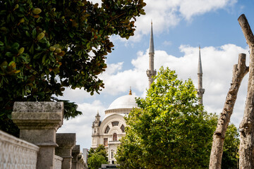 White mosque dome and tall minarets visible behind green tree branches on sunny day. Historic Islamic architecture and cultural heritage concept. © Tetiana