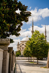View of historic Turkish mosque dome and minarets behind green trees under blue cloudy sky. Perspective shot of stone fence and courtyard conveys spiritual heritage and urban travel culture. © Tetiana