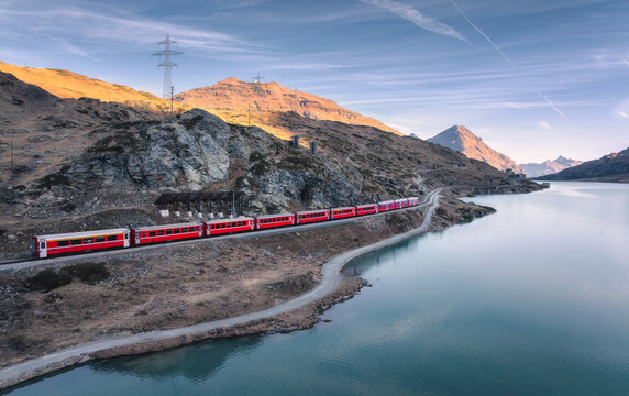 Aerial view of Bernina Express train near Bianco lake in the Swiss Alps at sunset, with glowing mountain peaks, water with reflections, rocky shore. Peaceful autumn landscape. Mountain train in fall