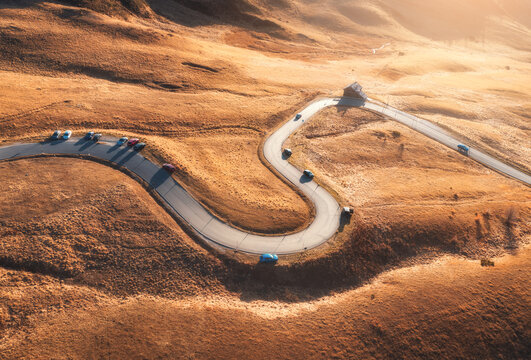 Winding mountain pass road with cars on orange grass slope in Dolomites, Italy. Aerial drone view of serpentine highway, autumn hills, hairpin turn, driving route and golden sunset light. Top view
