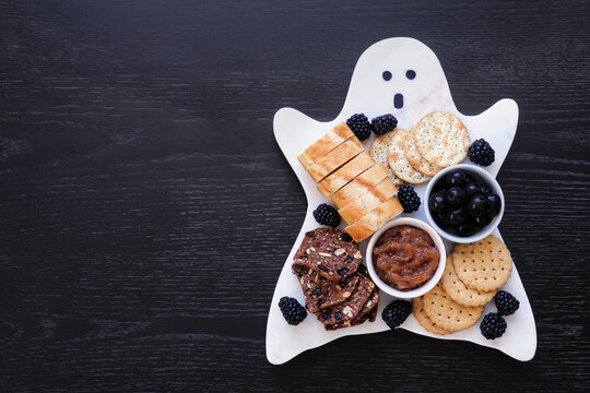 Spooky Halloween ghost charcuterie board with breads, blackberries, crackers, caramelized onions and olives. Top down view on a black wood background.