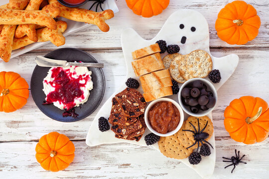 Spooky Halloween ghost charcuterie board with breads, blackberries, crackers, caramelized onions, olives and a side of bones and blood splatter cream cheese. Overhead view on a white wood background.
