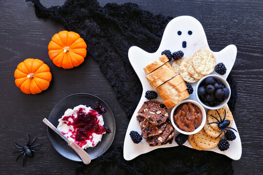 Spooky Halloween ghost charcuterie board with breads, blackberries, crackers, caramelized onions, olives and a side of blood splatter cream cheese. Top view table scene on a black wood background.