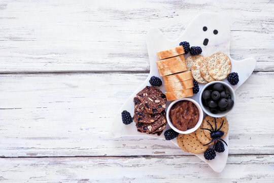 Spooky Halloween ghost charcuterie board with breads, blackberries, crackers, caramelized onions and olives. Overhead view on a white wood background.