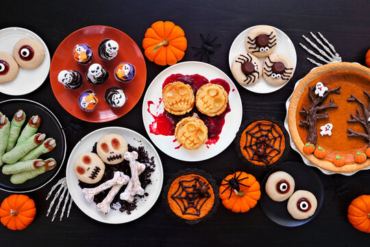 Fun Halloween dessert table scene over a black wood background. Overhead view. Variety of cookies, cupcakes and pumpkin and apple tarts and pies.