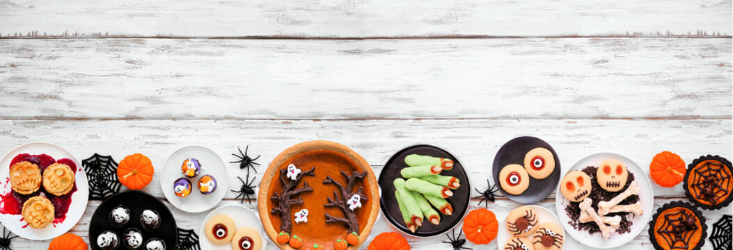 Fun Halloween dessert bottom border over a white wood banner background with copy space. Top down view.  Assortment of cookies, cupcakes and pumpkin and apple tarts and pies.