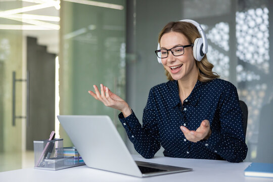 Businesswoman with glasses and wireless headset smiling during a virtual meeting on her laptop in a modern office, participating in remote work, webinar or online coaching session