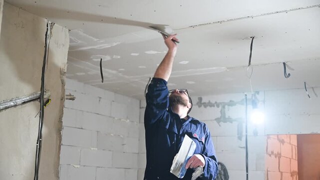Worker applies putty to ceiling during interior renovation. Builder smooths drywall joints and surface imperfections in unfinished room during finishing process