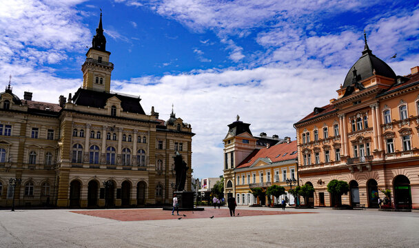 Pedestrians walk across Liberty Square (Trg Slobode) framed by the Neo-Renaissance City Hall and historic pastel buildings under a bright partly cloudy sky in Novi Sad, Serbia