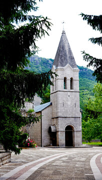 A stone bell tower and chapel rise from a paved courtyard framed by pine trees and lush green mountains at the historic Morača Monastery in Montenegro