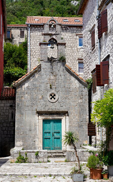 A small stone chapel featuring a turquoise wooden door and an open bell-gable stands in a paved courtyard surrounded by historic stone buildings in Kotor, Montenegro