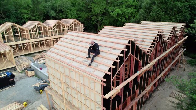 Designer on rooftop of wooden frame hospitality complex under development. Drone video shows resort buildings, pool section, and forest edge construction site