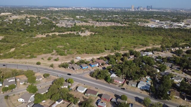 Paved arterial lanes curve past single-family homes across Historic Southside. Thick woodland corridors frame distant downtown high-rise silhouettes under clear skies, Fort Worth, Texas