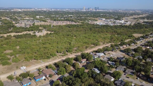 Unpaved perimeter road separates quiet residential backyards across DFW suburbs. Lush green forest canopy extends toward distant downtown skyline skyscrapers under sun, Fort Worth, Texas