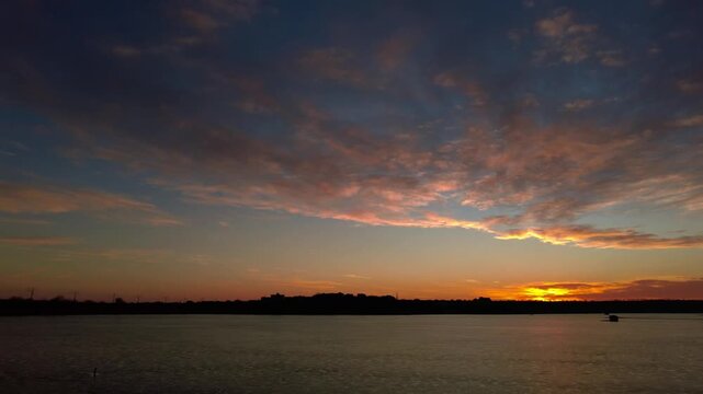 4K video of colorful sunset with dramatic clouds over frozen lake at dusk. Peaceful winter nature footage with silhouetted shoreline and reeds.
