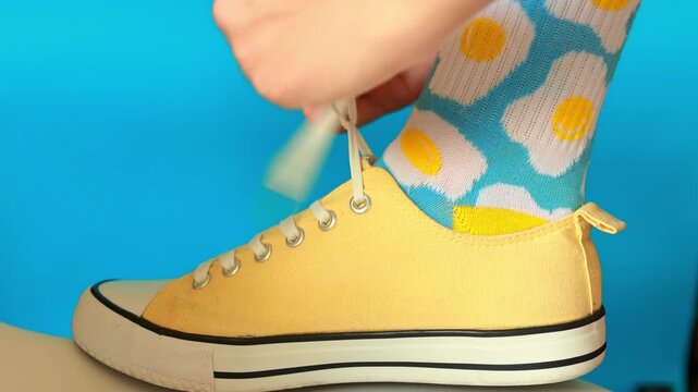 Close-up of a person tying the laces of bright yellow canvas sneakers while wearing light blue socks patterned with fried eggs and smiley faces against a vibrant blue background