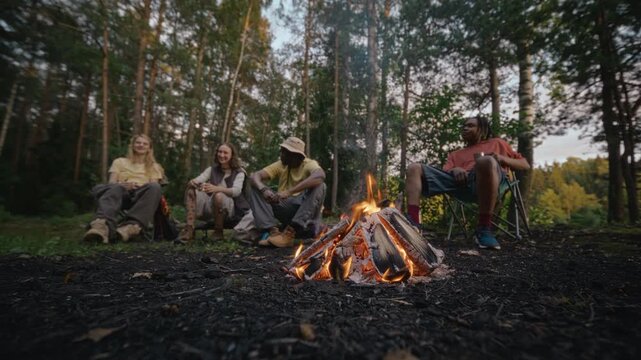 Diverse young adults sitting around campfire in forest, talking and relaxing during camping trip. Footage is suitable for outdoor gear brands or travel campaigns promoting adventure