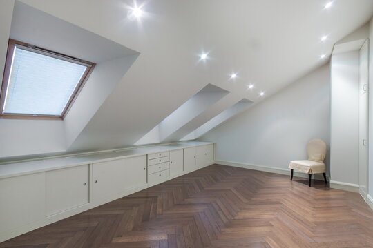 This space is an empty attic room featuring a wooden floor and an angled ceiling. There is a small chair and natural light coming through a window