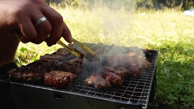A man turns cevapi with tongs on a smoky charcoal grill outdoors. Traditional barbecue footage suits food, travel, leisure and hospitality themes