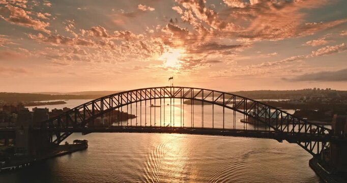 Sydney Harbour Bridge traffic arching over the water with the iconic Opera House nearby, capturing city skyline and dramatic orange clouds during a beautiful sunset. Aerial view drone flight panorama