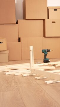 Cardboard boxes stack in the background. Drill rests on wooden planks. The floor shows natural wood grain. The scene suggests moving or home projects. No people present